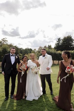Wedding party in brown and white attire poses playfully on a grassy lawn under cloudy skies.