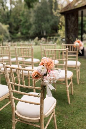 Garden wedding ceremony setup with white cushioned chairs decorated with coral flowers.