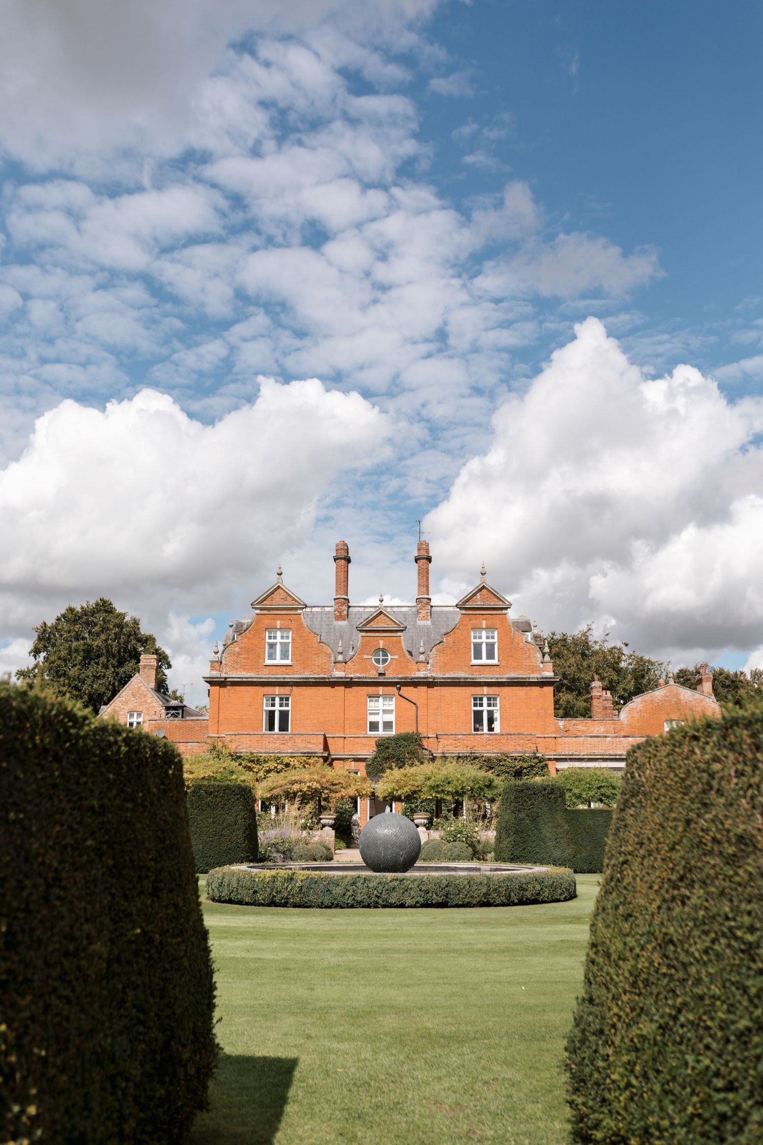 Stately red brick manor house with dormer windows set behind manicured lawn and hedges.