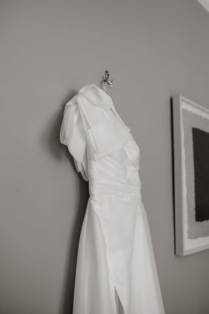 A white wedding dress hangs elegantly against a gray wall, showcasing its draped design and flowing fabric.