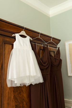 White flower girl dress hanging on wooden hangers next to brown bridesmaid dresses in a bedroom.
