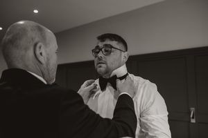 Someone helps adjust a black bowtie on a dress shirt in a dimly lit room.
