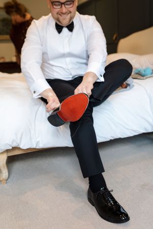 Person in formal attire sitting on white bedding holding a red table tennis paddle.
