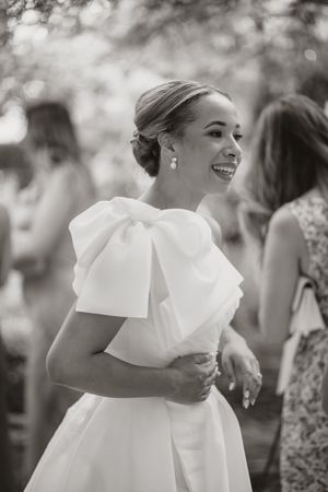 A bride in an elegant white wedding dress with a large bow detail smiles joyfully during an outdoor celebration.