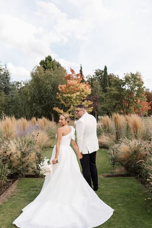 A couple in wedding attire twirls and dances in a garden setting with ornamental grasses and autumn foliage.