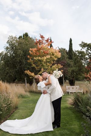 A romantic wedding couple embraces in a garden setting with autumn trees and ornamental grasses in the background.