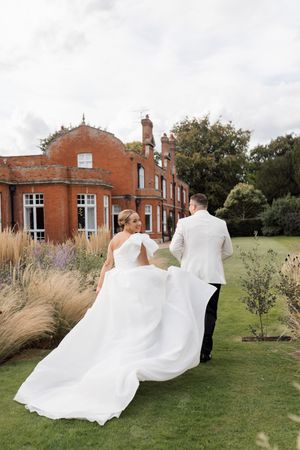 A flowing white wedding dress billows in the breeze near a red brick building with manicured grounds.