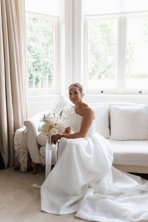 Elegant bride in white gown sits gracefully near sunlit windows holding a delicate bouquet of flowers.