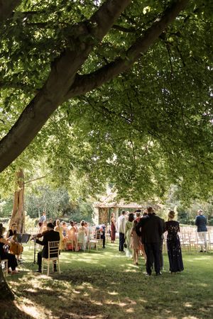 Wedding guests gather under large shade trees for an outdoor garden reception.