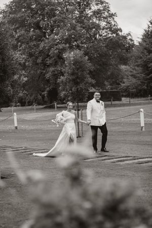 A couple in wedding attire walks together on a grassy path with trees in the background.