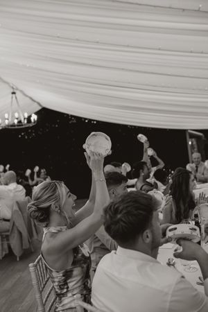 Black and white shot of wedding guests raising glasses in celebration under a white tent ceiling.