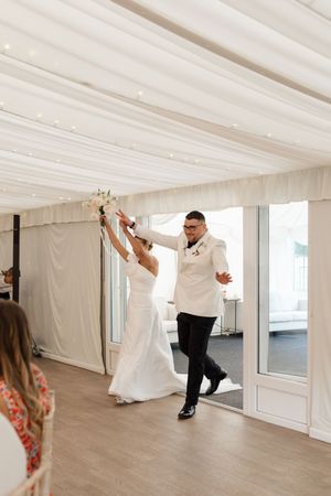 A couple dances playfully in an elegant white-draped reception tent with natural lighting.