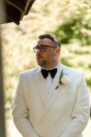 A groom in a crisp white tuxedo and black bow tie stands thoughtfully during an outdoor wedding ceremony.