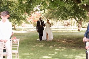Outdoor wedding ceremony setup with white chairs arranged on a lawn under shady trees on a sunny day.