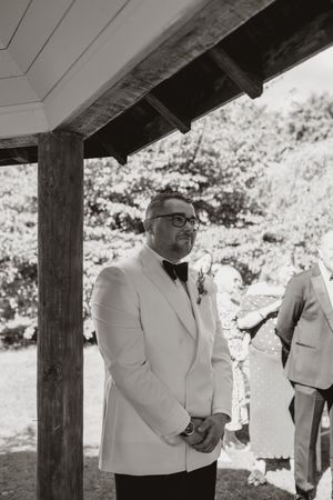Wedding ceremony guests in white jackets gathered under a wooden pergola outdoors.