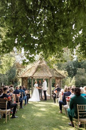 Outdoor wedding ceremony in progress under tree canopy with guests seated on chiavari chairs.