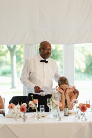A server in formal attire pours drinks for guests at an elegant wedding reception under a white tent.
