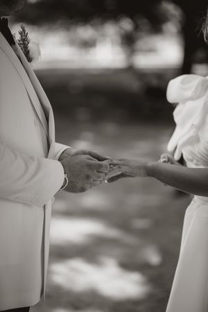 Black and white photo of formal-dressed people exchanging rings during outdoor ceremony.