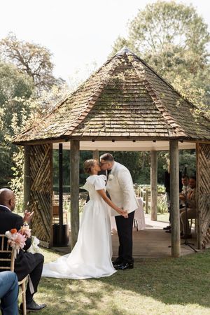 Wedding ceremony taking place under a wooden gazebo in a garden setting.