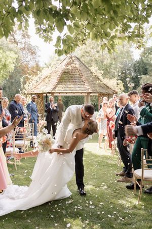 Newlyweds share romantic kiss while guests shower them with flower petals in garden gazebo setting.