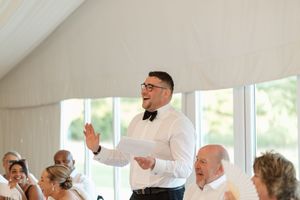 A guest delivers an animated wedding speech while standing among tables of seated guests in a bright reception tent.