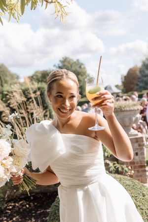 A person in a white wedding dress holds up a champagne glass while smiling outdoors in a garden setting.