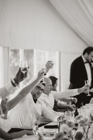 Wedding guests raise their glasses in celebration during a toast at an elegant reception under a white tent.