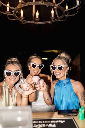 Friends wearing matching white cat-eye sunglasses pose together at a party with a rustic chandelier overhead.