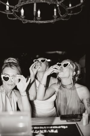 Three people wearing heart-shaped sunglasses laugh and pose together at a party in black and white photography.