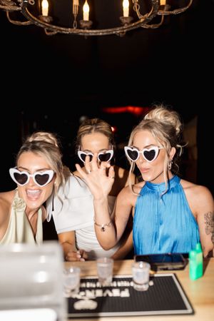 Group wearing heart-shaped sunglasses posing together at a party with drinks and dim lighting.