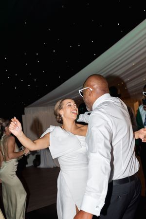 A couple dances together at a wedding reception under a white tent with twinkling lights overhead.