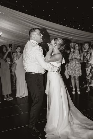 A couple dances together at a wedding reception under a white draped tent ceiling surrounded by guests.