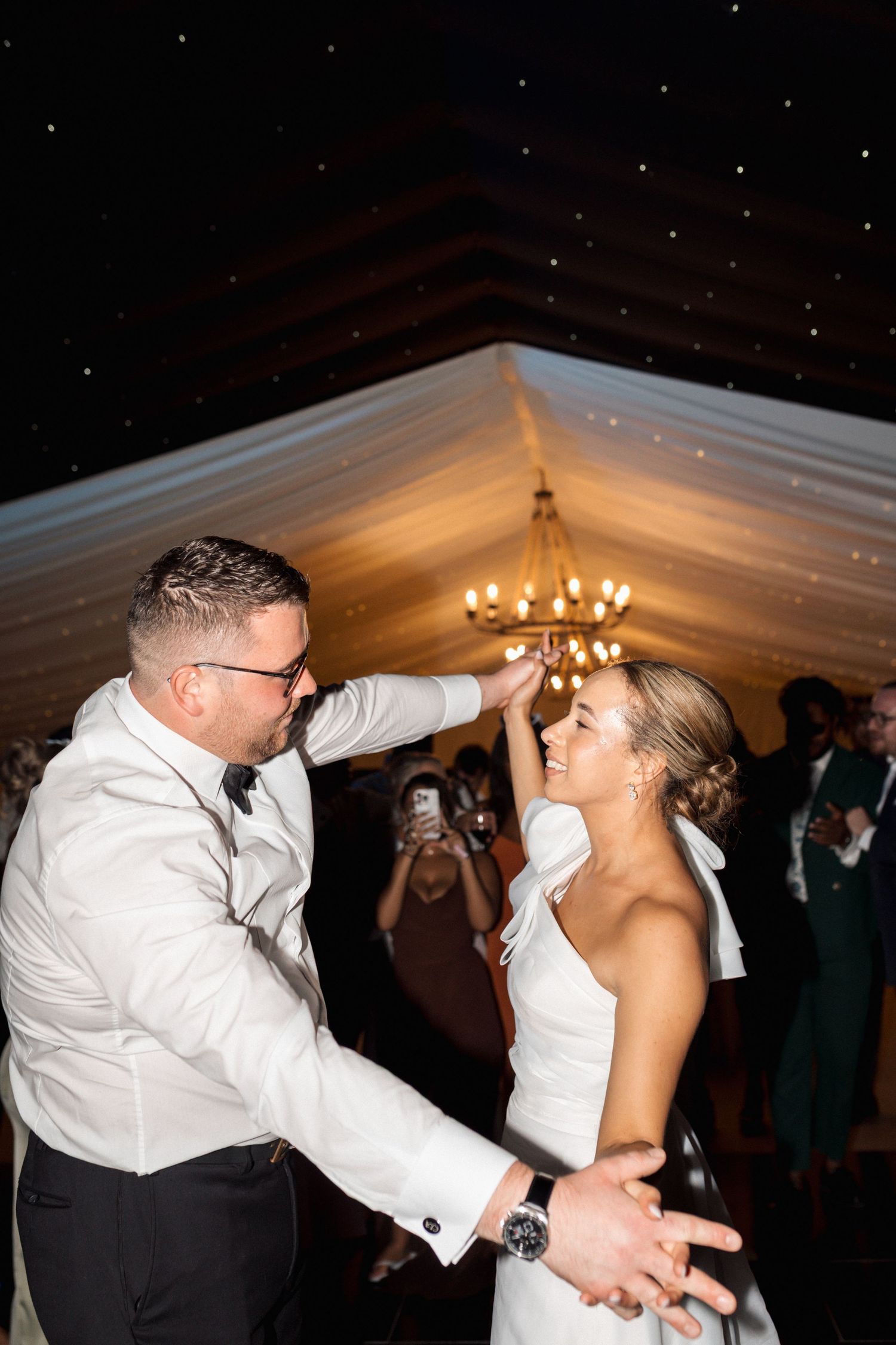 A couple shares a romantic dance dip moment in a white tent reception venue illuminated by warm chandelier lighting.