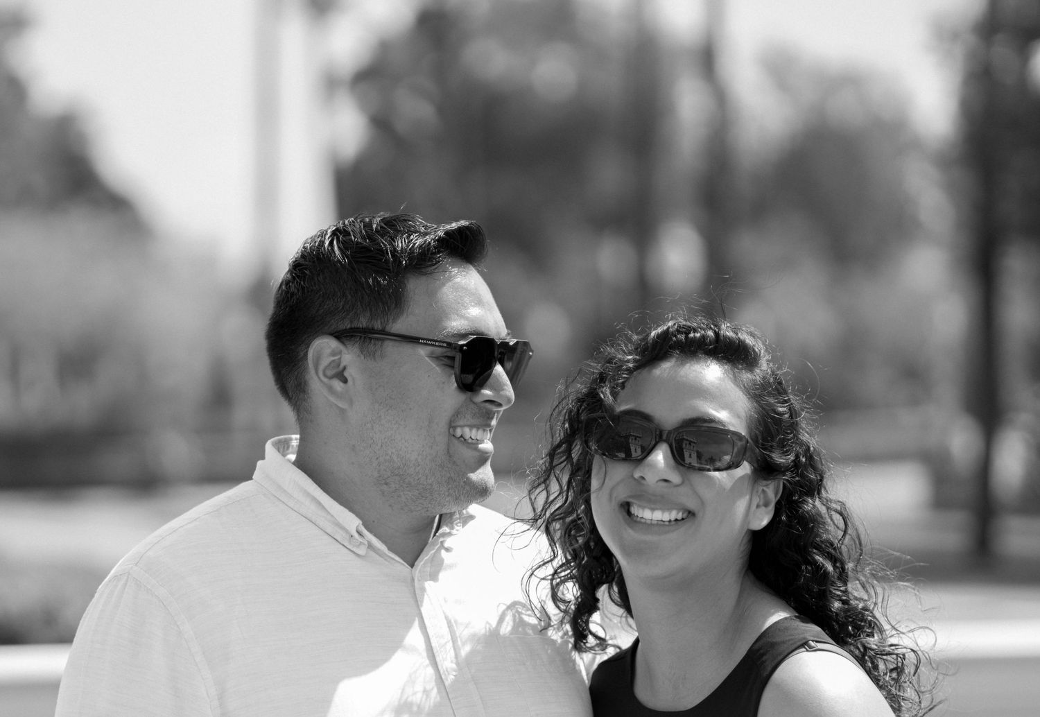 A black and white portrait of a smiling couple wearing sunglasses outdoors.