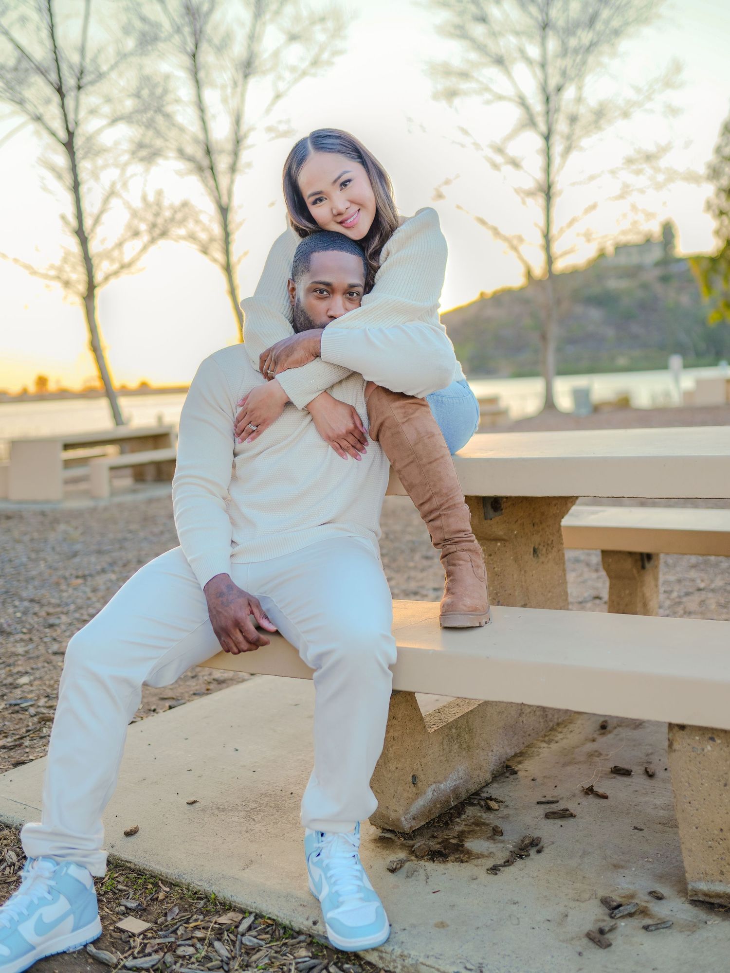 Casual outdoor photoshoot of couple in white outfits sitting on bench during sunset.