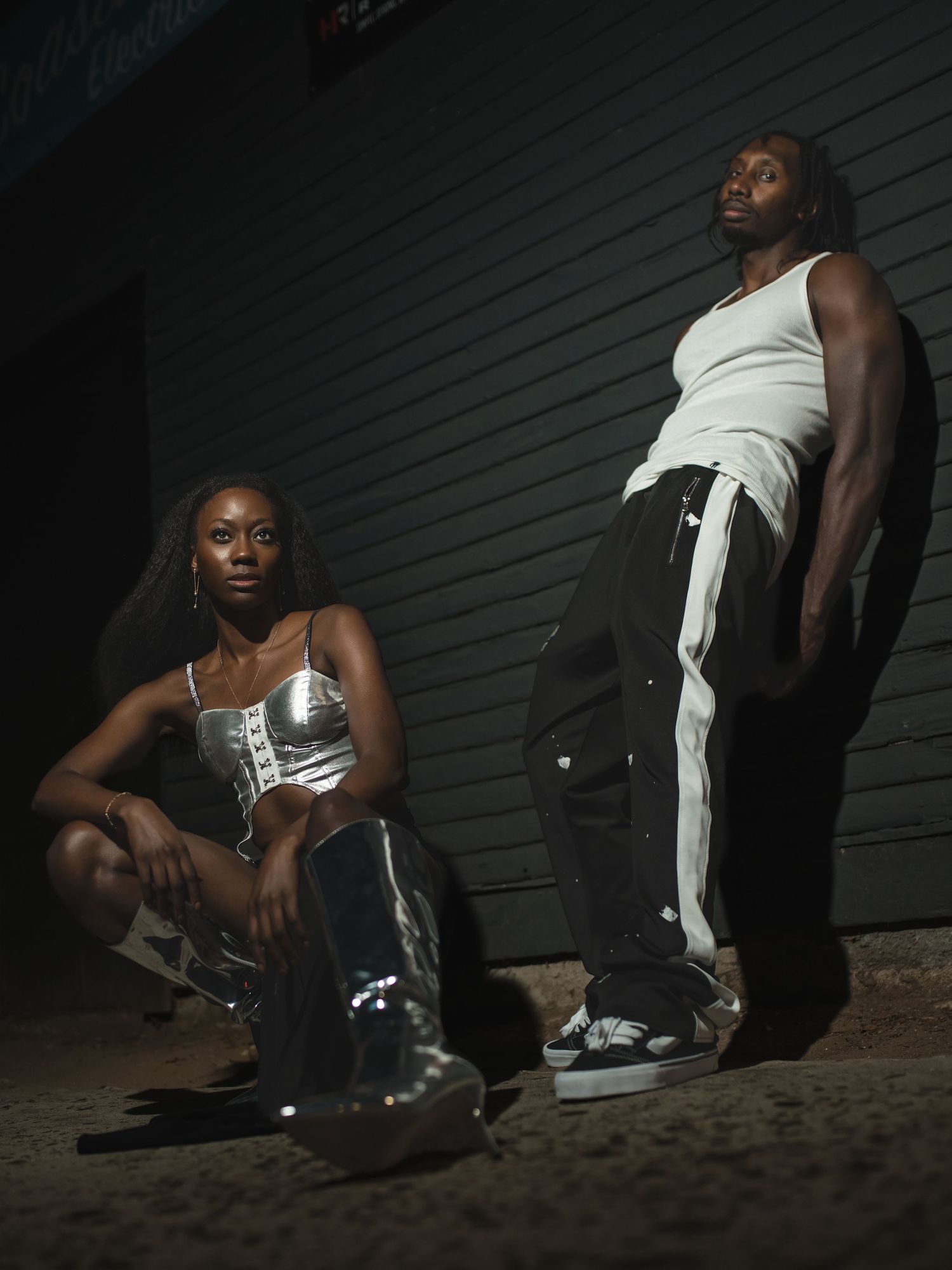 Urban fashion shoot featuring two models in monochromatic outfits with metallic details against dark wall.