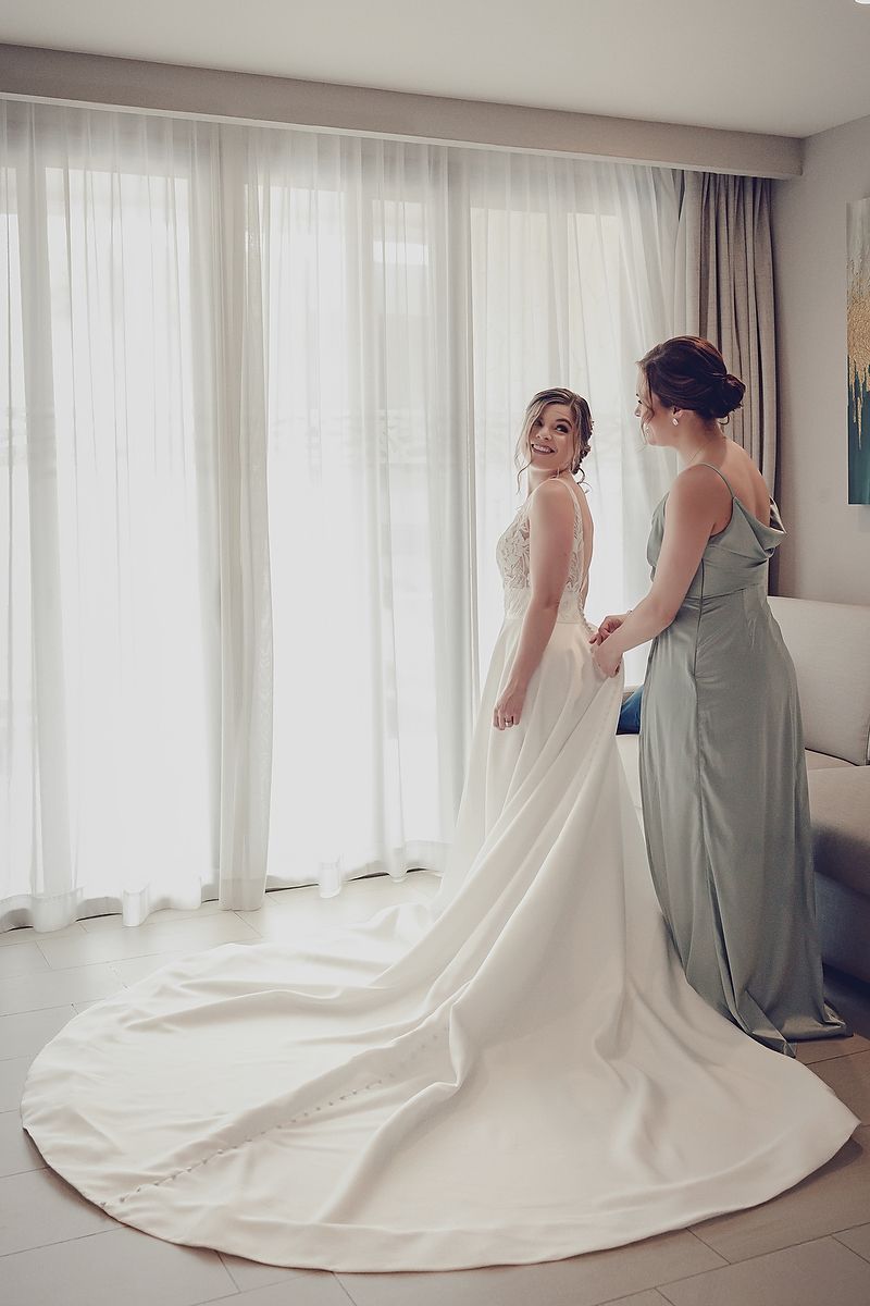 Two people stand near sunlit windows, one wearing a white wedding gown with a long train and another in a sage green dress.