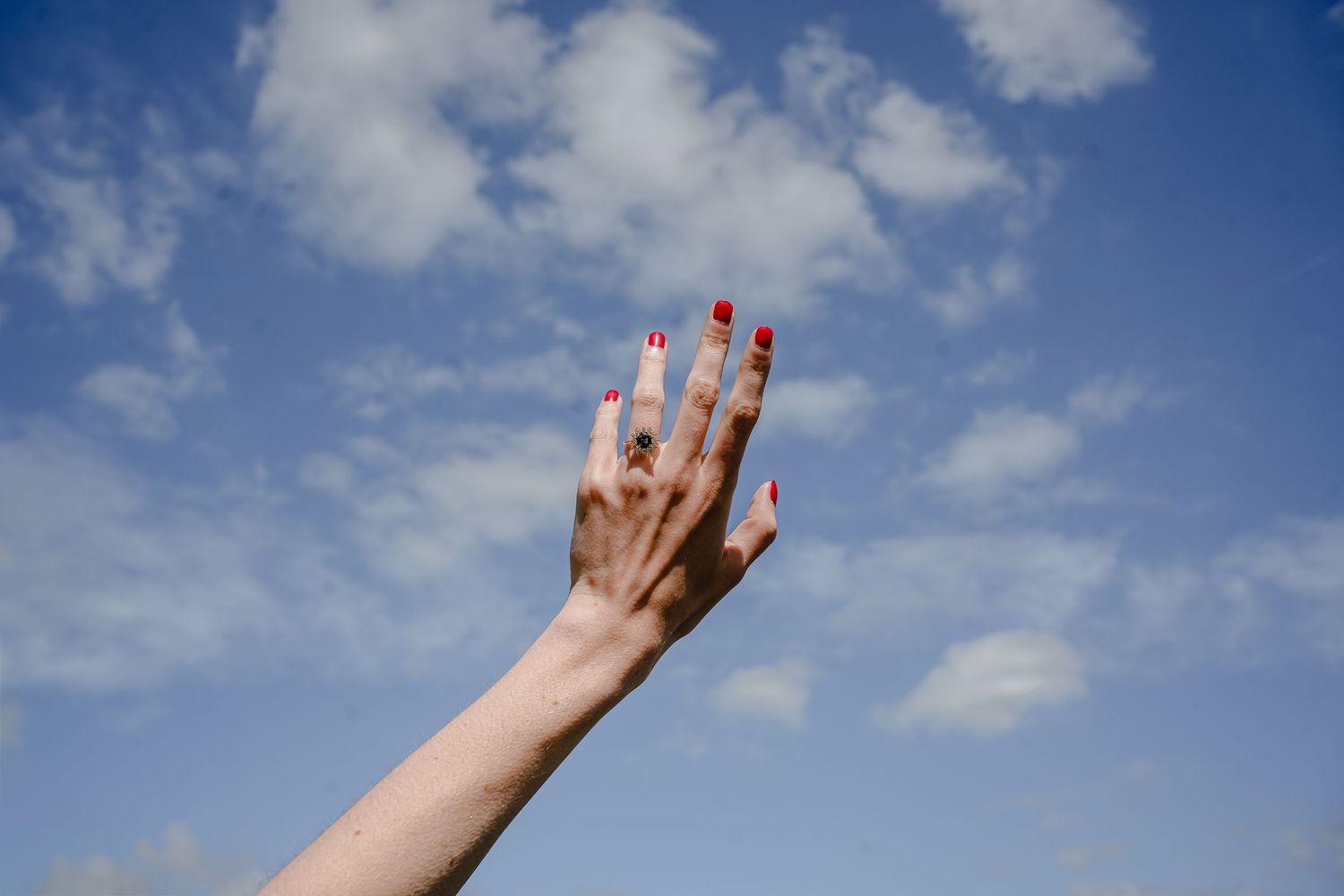 Hand with red nail polish reaches toward blue sky with fluffy white clouds.