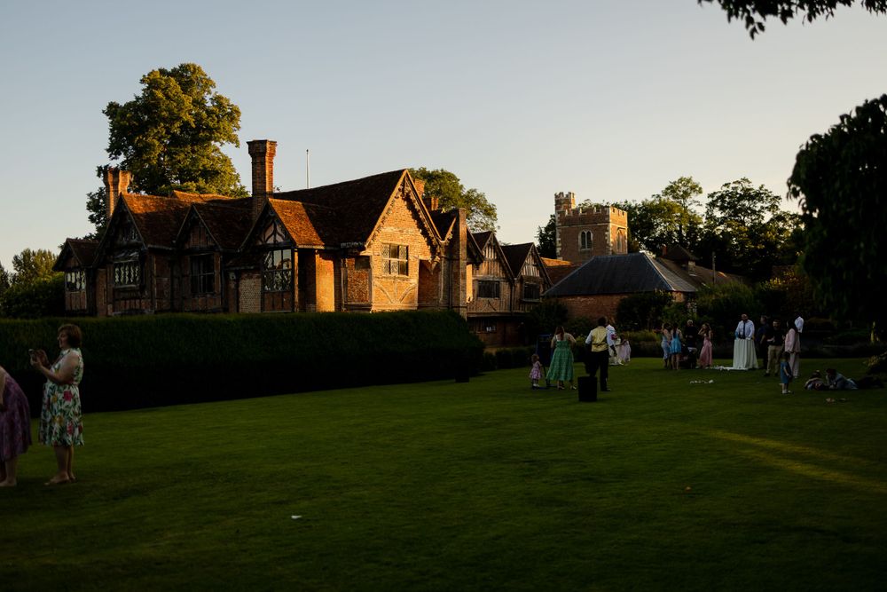 Wedding guests gather on a green lawn at sunset in front of a charming brick and timber English country house.