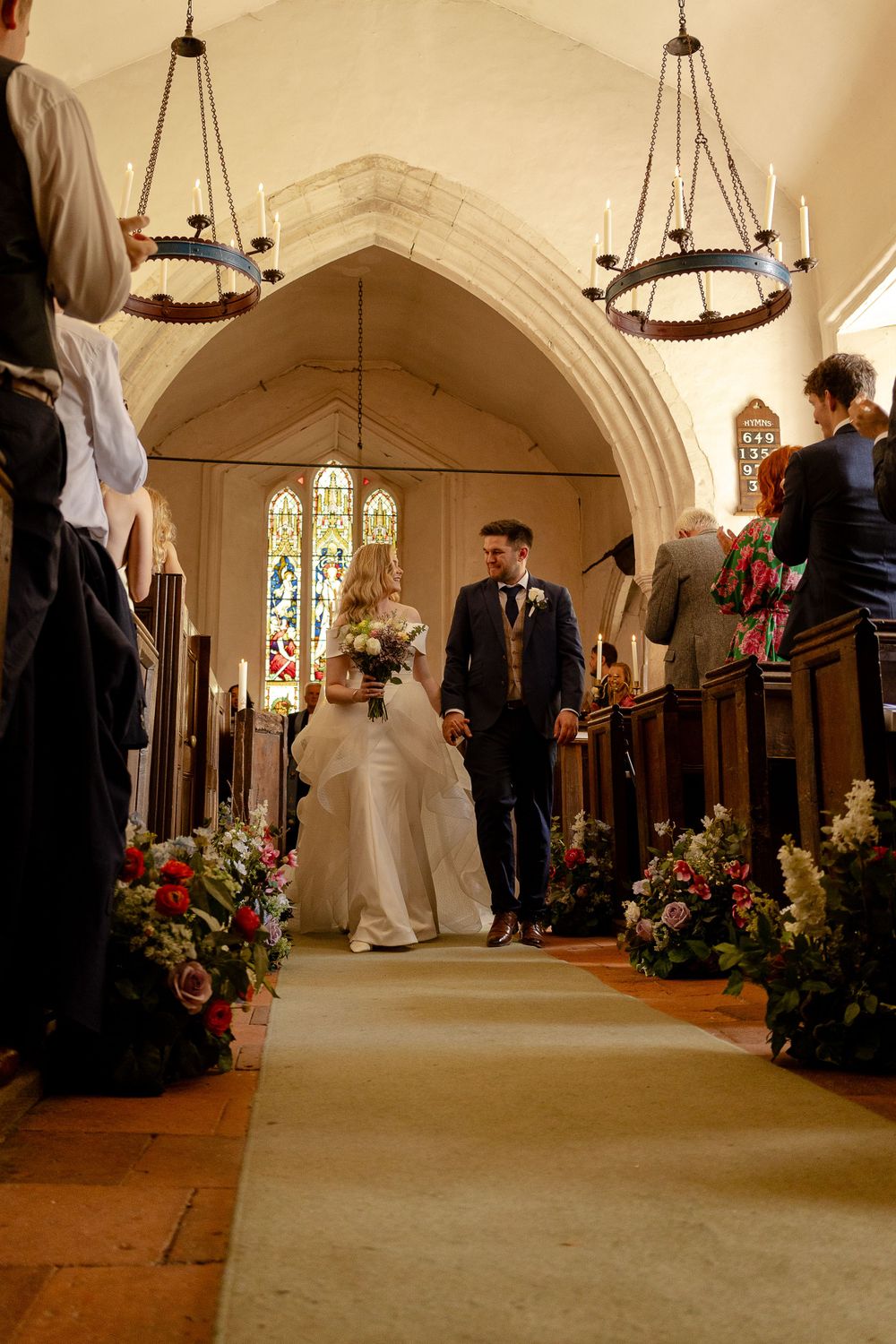 A wedding ceremony recessional in a traditional church with hanging chandeliers and stained glass windows.