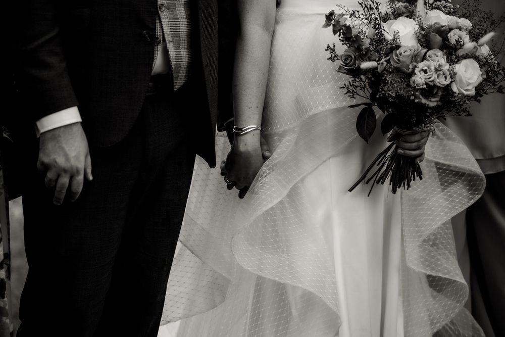 Close-up detail of wedding couple holding hands, showing dress and suit details in black and white.