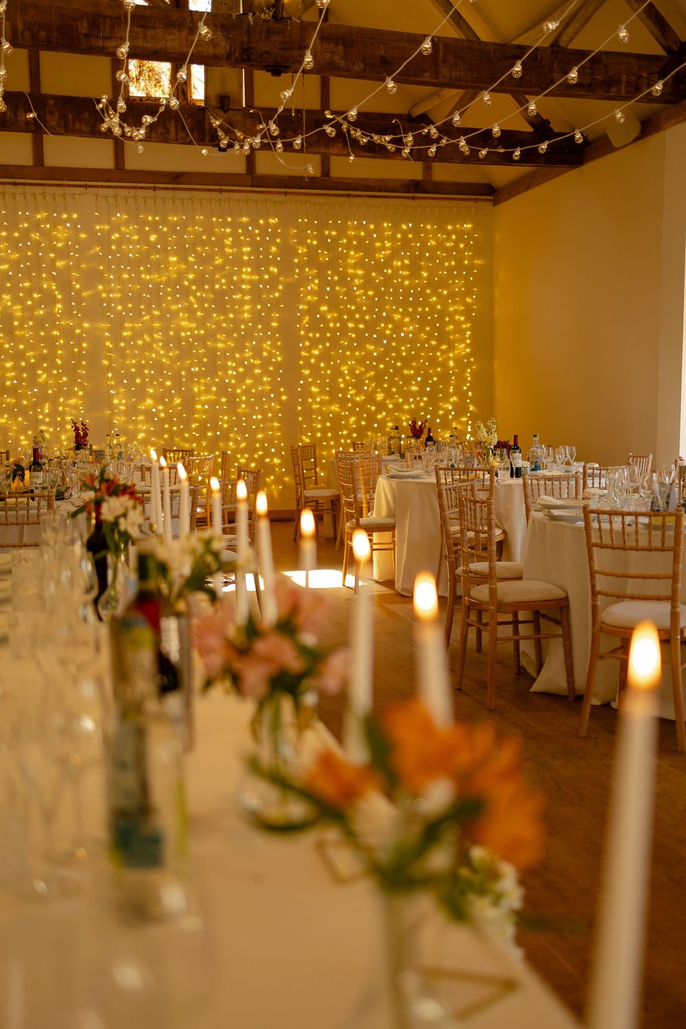 An elegant wedding reception venue with twinkle light backdrop, candles, and floral centerpieces on white-clothed tables.