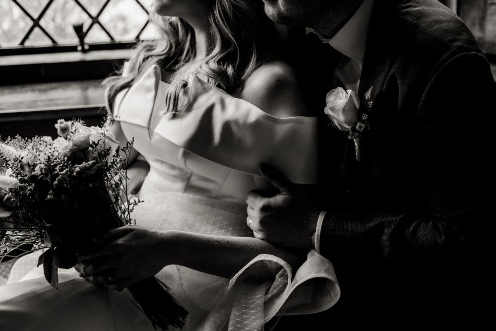 A black and white artistic shot of a romantic moment near a window with a wedding bouquet.