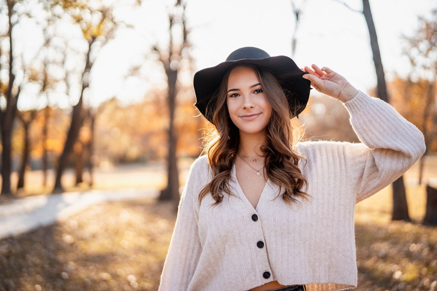 Woman in cream sweater and black hat poses outdoors during autumn with warm lighting.