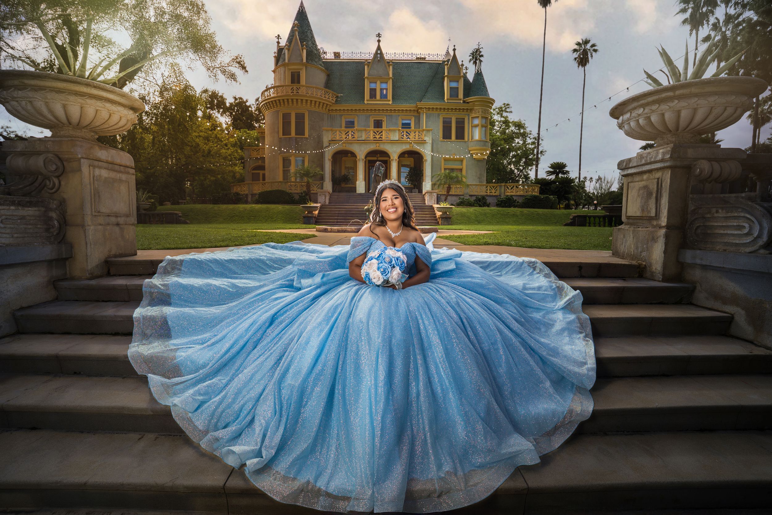 A figure in a flowing blue ball gown poses elegantly on stone steps at sunset for a photoshoot.