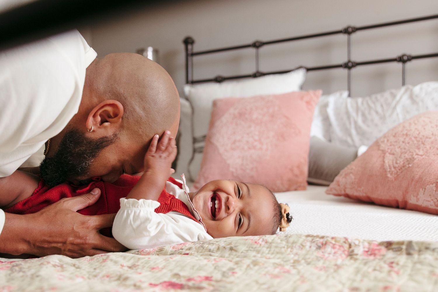 An infant with a hair bow rests peacefully on pink bedding while wearing a white onesie while dad tickles her belly