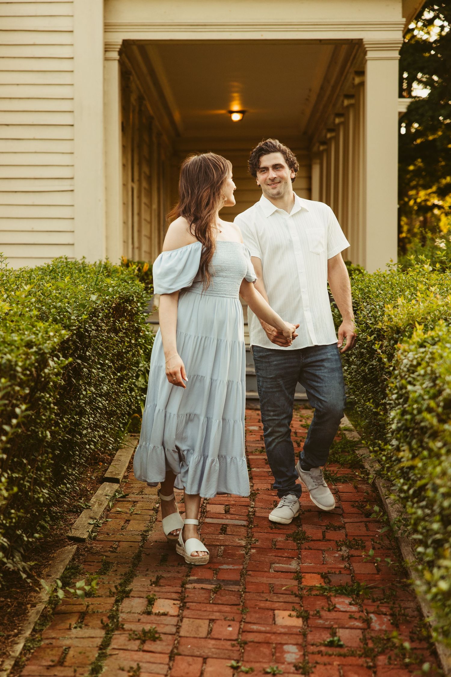 A couple stands together on a brick path with white columns and greenery, wearing casual summer outfits.