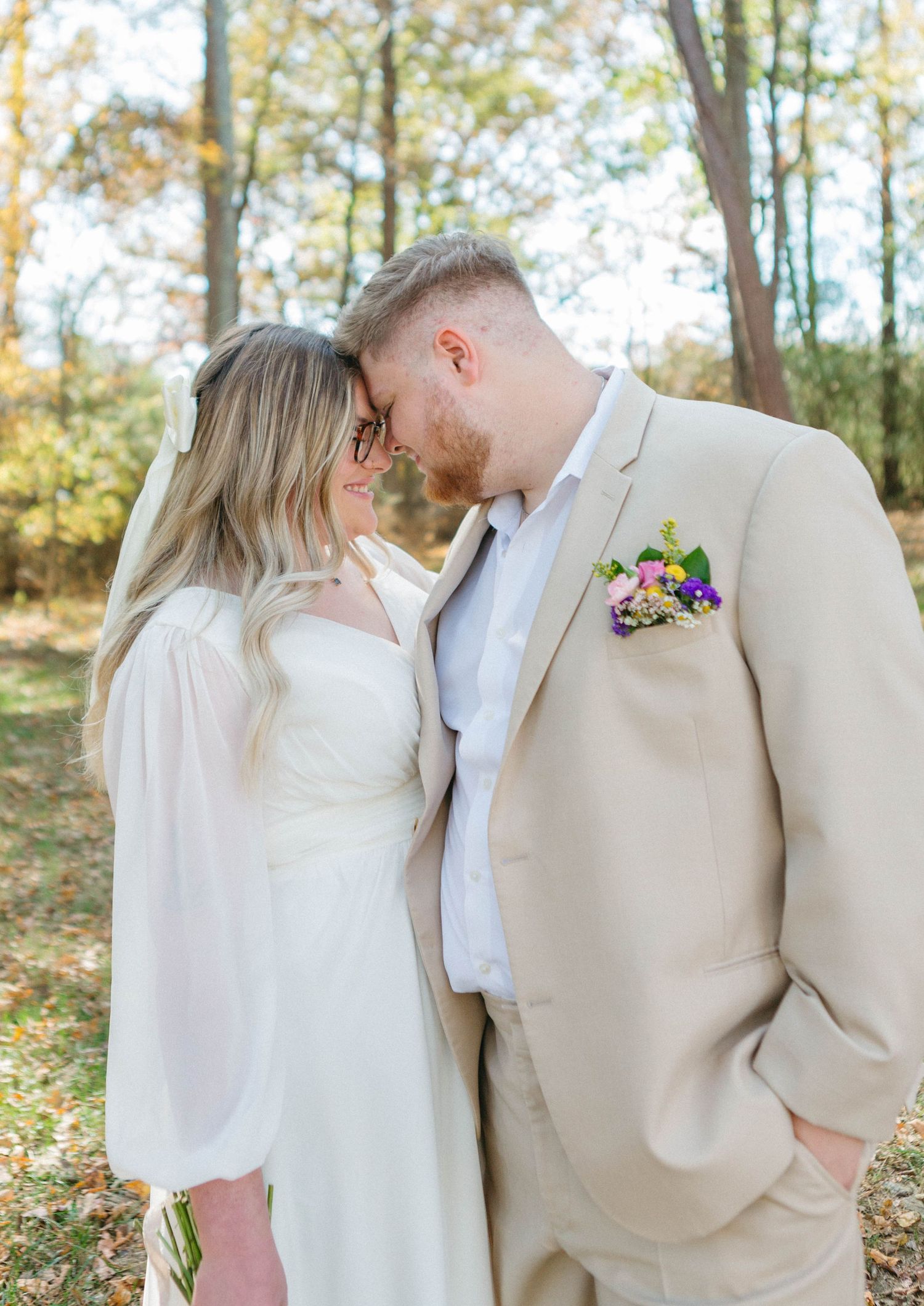 Newlywed couple in cream-colored attire sharing intimate moment in autumn forest setting.