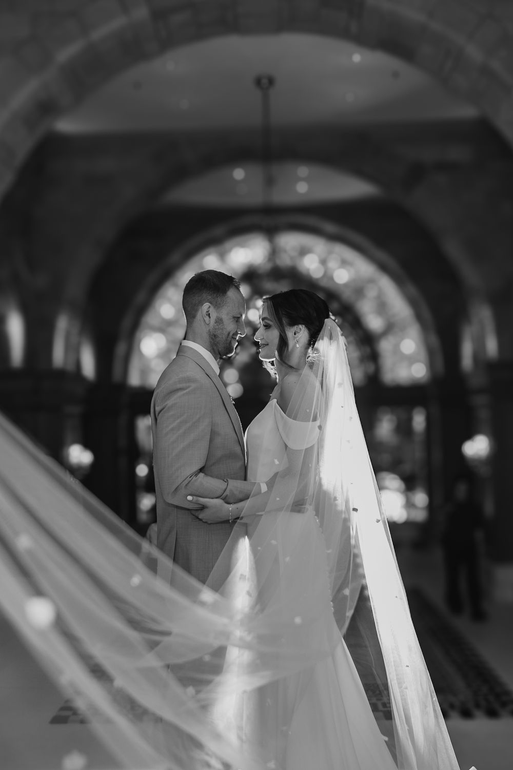 A romantic black and white wedding photo of a couple embracing under an arched hallway with a flowing veil.