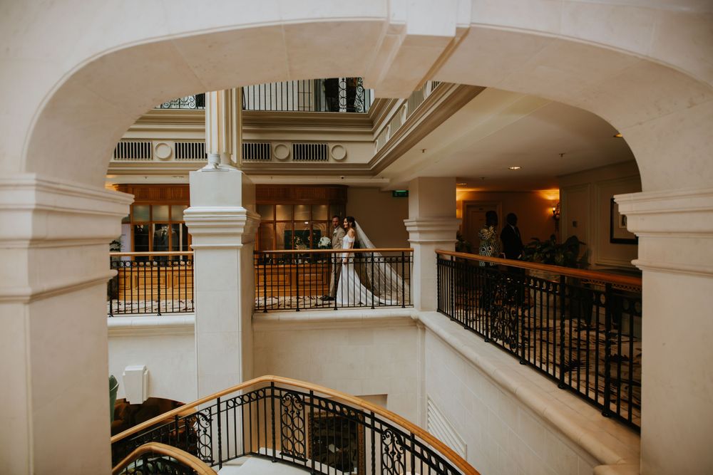 A bride stands at the top of an elegant curved staircase with ornate black railings and white pillars in a hotel interior.